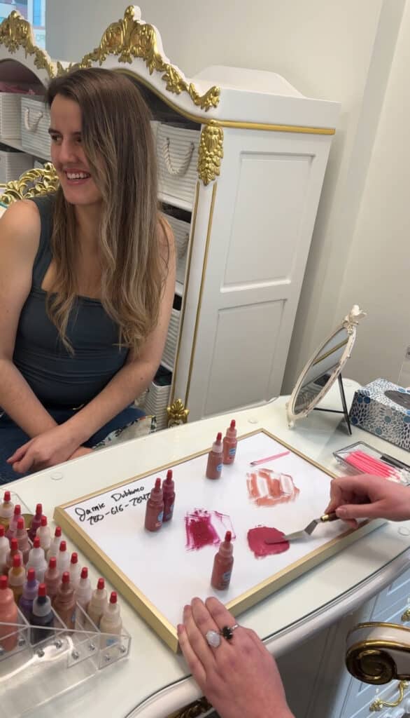 woman sitting at desk looking at a colour artist mix a custom blended mate liquid lipstick a Crème de la Crème Beauty Lounge